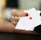 A hand with red nail polish holds two playing cards, including an ace with a diamond suit, against a blurred background.