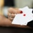 A hand with red nail polish holds two playing cards, including an ace with a diamond suit, against a blurred background.