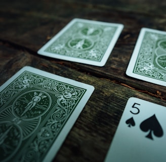 Several playing cards are scattered across a rustic wooden surface. The cards have intricate green designs on their backs, with one card facing up displaying the number five of spades.
