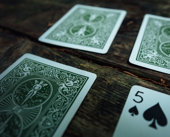 Several playing cards are scattered across a rustic wooden surface. The cards have intricate green designs on their backs, with one card facing up displaying the number five of spades.