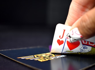 A hand is holding two playing cards, the Jack of Hearts and the Queen of Hearts, revealing part of the hand in a game of poker. The cards are partially lifted from a branded poker deck displaying a logo. The background is dark, emphasizing the cards and hand in focus.