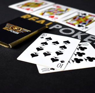 A deck of playing cards labeled 'Real Poker' rests on a dark surface. Two cards, the ten of spades and ten of clubs, are prominently displayed in the foreground. A row of face cards, including kings and queens from different suits, is lined up in the background.