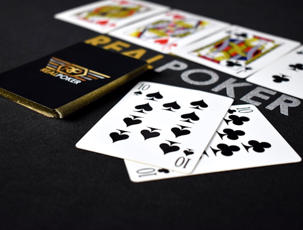A deck of playing cards labeled 'Real Poker' rests on a dark surface. Two cards, the ten of spades and ten of clubs, are prominently displayed in the foreground. A row of face cards, including kings and queens from different suits, is lined up in the background.