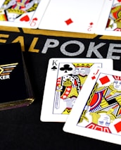 Poker-themed setup featuring a deck of cards on a table with the words 'REAL POKER' visible. Two cards, the King of Clubs and Jack of Diamonds, are prominently displayed. The design is vibrant, with detailed illustrations on the cards.
