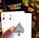 A hand holding two playing cards, an Ace of Clubs and an Ace of Spades, with a backdrop of gold Bitcoin tokens. The hand has an orange fingernail with a white Bitcoin symbol on it. The background features a stack of poker chips with a logo that reads 'Real Poker'.