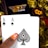 A hand holding two playing cards, an Ace of Clubs and an Ace of Spades, with a backdrop of gold Bitcoin tokens. The hand has an orange fingernail with a white Bitcoin symbol on it. The background features a stack of poker chips with a logo that reads 'Real Poker'.