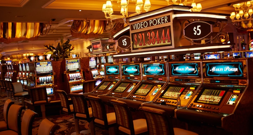 A casino floor featuring rows of brightly lit video poker machines. The area is illuminated by elegant chandeliers, with a display board showing a large sum of money, suggesting a possible jackpot. The rich decor includes luxurious carpets and comfortable seating, adding to the opulent atmosphere.