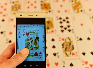 A hand is holding a smartphone displaying a close-up image of playing cards, specifically a King of Hearts and an Ace of Clubs. In the background, several playing cards are spread out, featuring various suits and numbers, slightly blurred for depth of field.