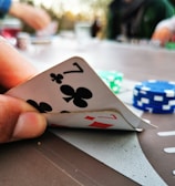 A close-up of a hand holding two playing cards, a seven of clubs and a seven of diamonds. In the background, there are poker chips in different colors, such as blue and green, scattered on a table. The image suggests a casual setting with people playing poker.