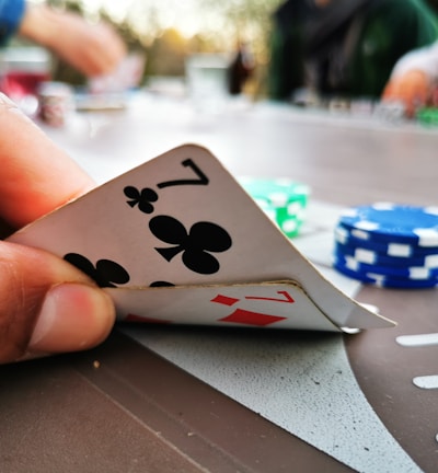 A close-up of a hand holding two playing cards, a seven of clubs and a seven of diamonds. In the background, there are poker chips in different colors, such as blue and green, scattered on a table. The image suggests a casual setting with people playing poker.