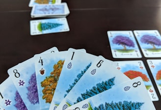 A hand holds several playing cards with tree illustrations and different numbers, set against a dark table. Additional cards are arranged in rows on the table, showing various tree images and numbers.