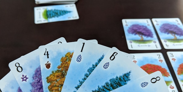 A hand holds several playing cards with tree illustrations and different numbers, set against a dark table. Additional cards are arranged in rows on the table, showing various tree images and numbers.