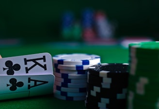 A casino scene featuring two playing cards, the ace and king of clubs, partially concealed by a player's hand. Nearby, there are stacks of multicolored poker chips on a green felt table, with blurred colorful background elements suggesting other chip stacks or cards.