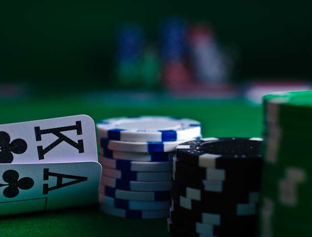A casino scene featuring two playing cards, the ace and king of clubs, partially concealed by a player's hand. Nearby, there are stacks of multicolored poker chips on a green felt table, with blurred colorful background elements suggesting other chip stacks or cards.