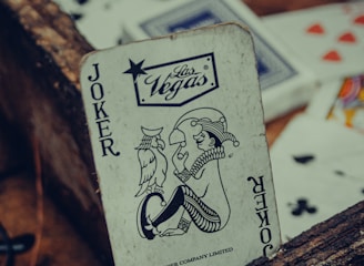 A playing card featuring a joker with a Las Vegas logo is prominently positioned among other scattered cards. The card has a vintage, worn look, resting against a rustic wooden surface. Various playing cards, partially visible, are scattered around with visible suits such as hearts and clubs.