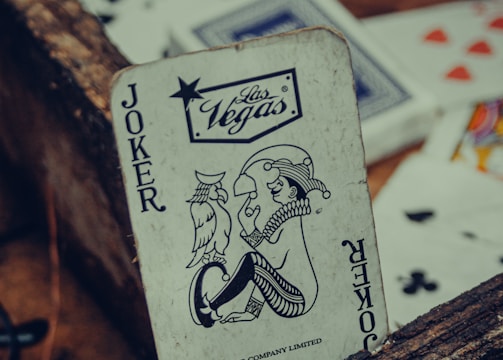 A playing card featuring a joker with a Las Vegas logo is prominently positioned among other scattered cards. The card has a vintage, worn look, resting against a rustic wooden surface. Various playing cards, partially visible, are scattered around with visible suits such as hearts and clubs.