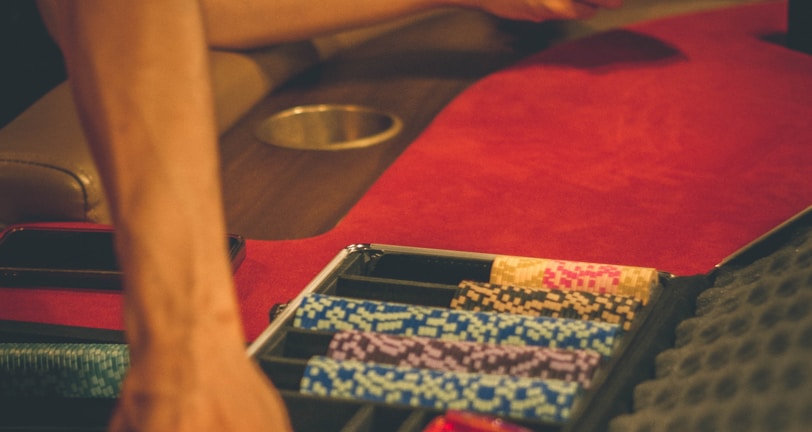 A person reaches into a poker chip case on a red felt table, while another person holds playing cards. The scene is dimly lit, creating a warm and engaged atmosphere.
