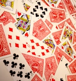 A vibrant assortment of playing cards scattered in a chaotic manner. The cards display various suits and numbers, predominantly featuring the colors red and black with intricate designs. Several face cards are visible, showcasing traditional royal characters in vivid detail.