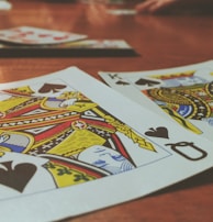 Playing cards are spread out on a wooden table, with the Queen and King of Spades prominently visible. The focus is on the vibrant colors and detailed designs of the cards, while other cards and hands are slightly blurred in the background, suggesting a game in progress.
