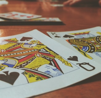 Playing cards are spread out on a wooden table, with the Queen and King of Spades prominently visible. The focus is on the vibrant colors and detailed designs of the cards, while other cards and hands are slightly blurred in the background, suggesting a game in progress.