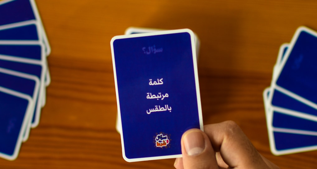 A hand holds a playing card with Arabic text printed on it. Several other cards are spread out on a wooden surface in different positions.