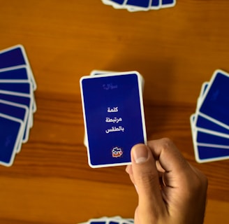 A hand holds a playing card with Arabic text printed on it. Several other cards are spread out on a wooden surface in different positions.
