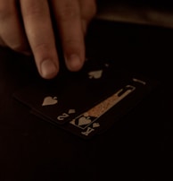 A hand is touching a stack of playing cards laid out on a dark surface. The cards are primarily black, featuring heart suits, including the king and jack. The scene is dimly lit, giving it an intimate or mysterious atmosphere.