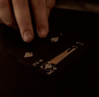 A hand is touching a stack of playing cards laid out on a dark surface. The cards are primarily black, featuring heart suits, including the king and jack. The scene is dimly lit, giving it an intimate or mysterious atmosphere.