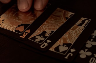 A hand gently resting on a spread of playing cards, featuring black suits and gold, ornate designs. The setting appears to be dimly lit, giving off a sophisticated and mysterious atmosphere.