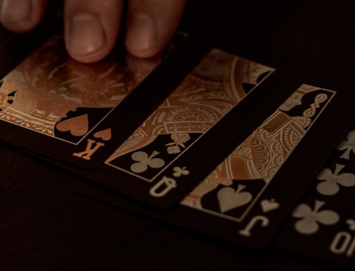 A hand gently resting on a spread of playing cards, featuring black suits and gold, ornate designs. The setting appears to be dimly lit, giving off a sophisticated and mysterious atmosphere.