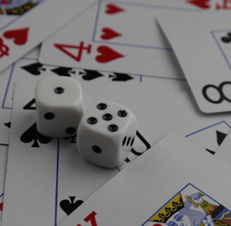 A collection of playing cards spread out, featuring various suits such as hearts, clubs, and spades. Two white dice with black dots are placed prominently on top of the cards.