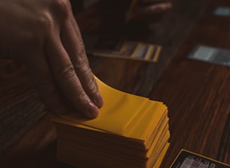 A hand is holding a stack of playing cards with yellow sleeves on a wooden table. Several cards are laid out in the background, and a dice is visible to the side.