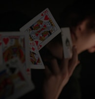 A person holding a playing card in their hand, with other playing cards floating in mid-air. The background is dark, highlighting the contrast between the cards and the surroundings.