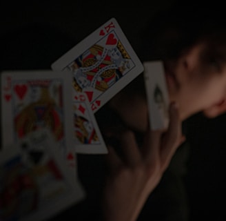 A person holding a playing card in their hand, with other playing cards floating in mid-air. The background is dark, highlighting the contrast between the cards and the surroundings.