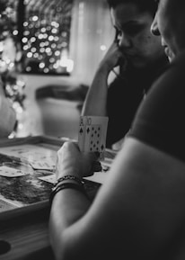 Three people are engaged in a card game, sitting around a table in a cozy, dimly lit setting. One player is holding an Ace and a Ten of diamonds. There are fairy lights and a blurred Christmas tree in the background, adding a festive and warm atmosphere to the scene.