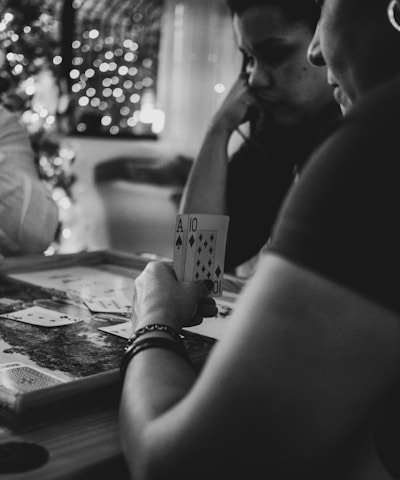 Three people are engaged in a card game, sitting around a table in a cozy, dimly lit setting. One player is holding an Ace and a Ten of diamonds. There are fairy lights and a blurred Christmas tree in the background, adding a festive and warm atmosphere to the scene.